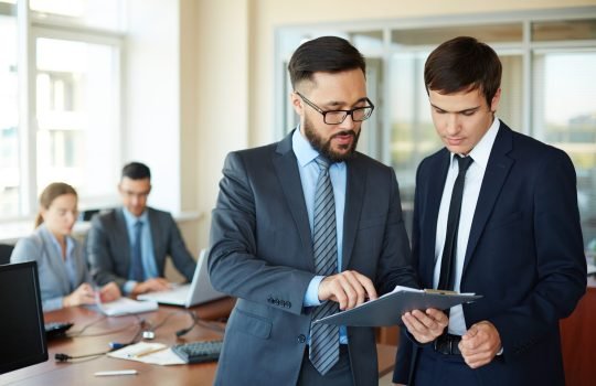 Confident businessman pointing at document while explaining his idea to his partner on background of their colleagues